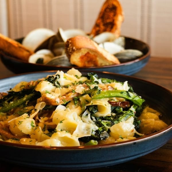 Pasta with greens and cheese in a dark bowl, with a bowl of clams and bread in the background.