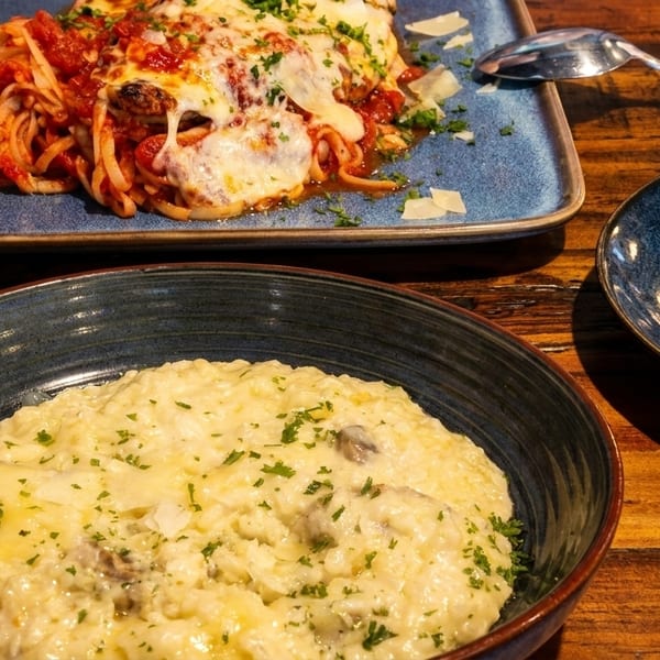 A bowl of creamy mushroom risotto and a plate of cheesy baked spaghetti with meatballs on a wooden table.